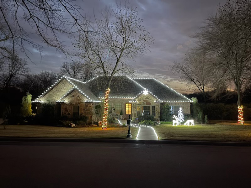 Brick ranch home at dusk with white lights on roofline and lighted reindeer in yard